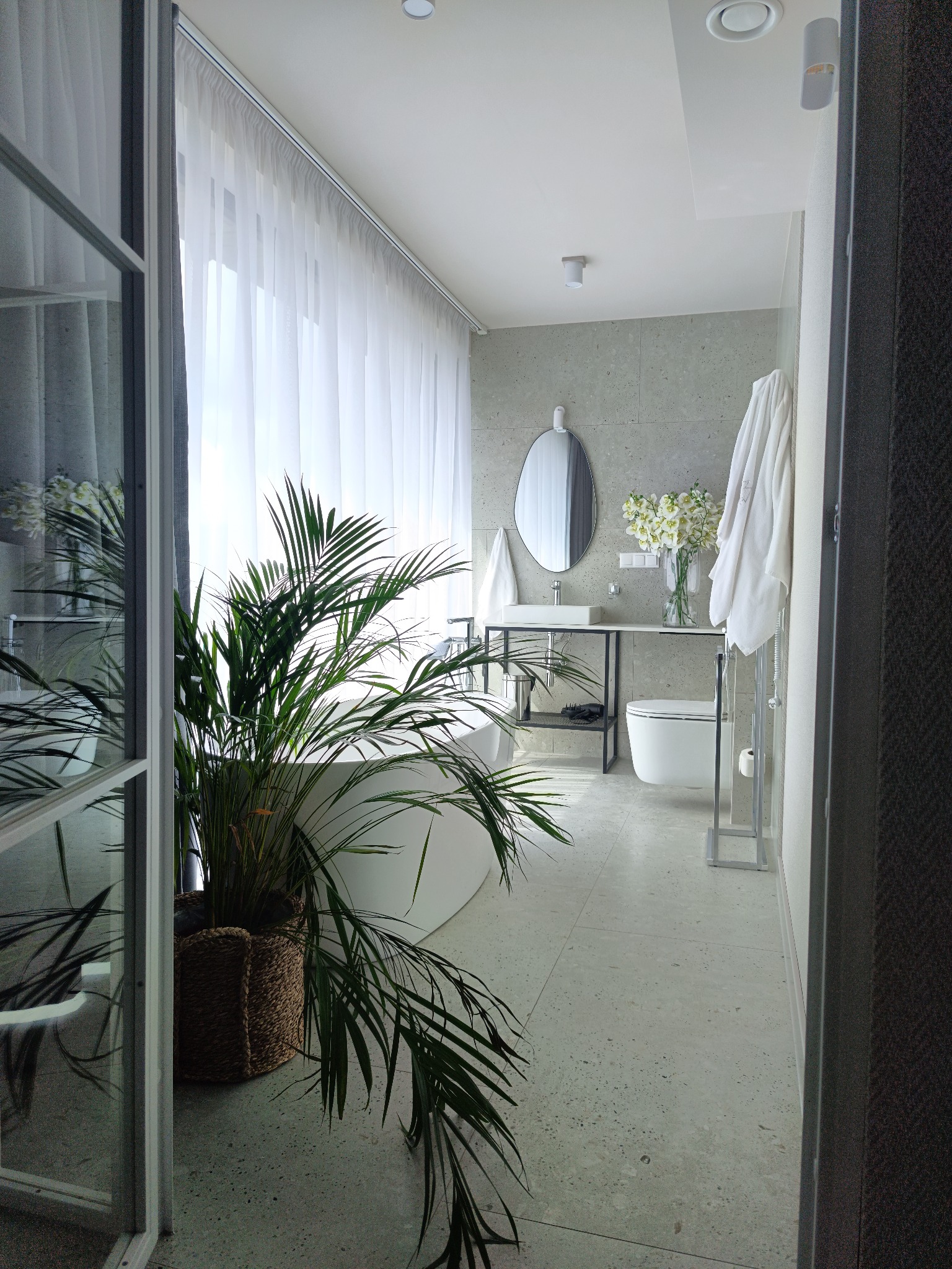 Jasna łazienka z wanną, umywalką na metalowym stelażu, toaletą i rośliną w koszu. Minimalistyczny design z szarymi płytkami i białymi zasłonami.