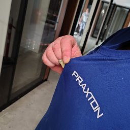 PRAXTON Spółka z o.o. - Montaż okien z bliska: dłoń trzyma klin, w tle widoczne zamontowane okna z ciemnymi ramami, odbijające światło. Na pierwszym planie fragment niebieskiej koszulki z napisem.