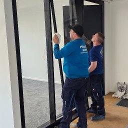 PRAXTON Spółka z o.o. - Montaż czarnej ramy okiennej w biurze: dwóch monterów w niebieskich uniformach instaluje okno, w tle widoczna pusta przestrzeń biurowa i podwieszany sufit.