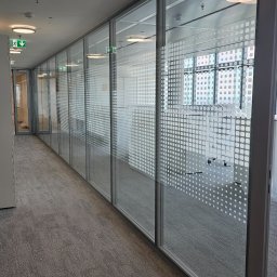 PRAXTON Spółka z o.o. - Szklane ściany działowe z dekoracyjną folią w biurze, widok z perspektywy korytarza. Minimalistyczny design, szara wykładzina. Oświetlenie sufitowe.