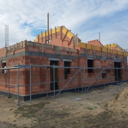 PRAXTON Spółka z o.o. - Budowa domu z czerwonej cegły, widoczne rusztowania i elementy konstrukcji dachu. Częściowo wzniesione ściany i przygotowane otwory okienne.