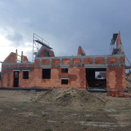 PRAXTON Spółka z o.o. - Budowa domu jednorodzinnego w trakcie wznoszenia ścian z czerwonej cegły. Widoczne rusztowania i przygotowany teren. Pochmurne niebo w tle, sąsiednie budynki.