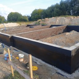 PRAXTON Spółka z o.o. - Zabezpieczone fundamenty domu z bloczków betonowych, wypełnione piaskiem, z widocznymi narzędziami budowlanymi i otoczeniem placu budowy w słoneczny dzień.