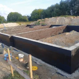 Zabezpieczone fundamenty domu z bloczków betonowych, wypełnione piaskiem, z widocznymi narzędziami budowlanymi i otoczeniem placu budowy w słoneczny dzień.