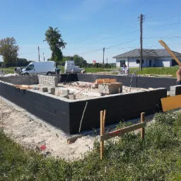 Wznoszenie fundamentów domu jednorodzinnego: murowane ściany z izolacją, pracownicy w trakcie robót, widoczny sprzęt budowlany i biały van w tle.