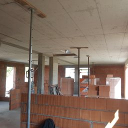 PRAXTON Spółka z o.o. - Wnętrze domu w trakcie budowy: ściany z czerwonej cegły, betonowy strop podparty metalowymi stemplami, okna bez ram. Widoczne materiały budowlane.