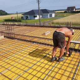 PRAXTON Spółka z o.o. - Prace zbrojarskie na budowie domu jednorodzinnego: zbrojarz łączy stalowe pręty na żółtym szalunku, w tle dom z ciemną dachówką i krajobraz wiejski.