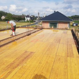 PRAXTON Spółka z o.o. - Przygotowanie szalunku i zbrojenia pod fundament domu jednorodzinnego. Widoczny pracownik impregnujący drewniane płyty szalunkowe. W tle zabudowa jednorodzinna i pola uprawne.