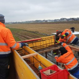 PRAXTON Spółka z o.o. - Trzech pracowników w pomarańczowych kurtkach montuje zbrojenie w drewnianym szalunku na tle pola i zabudowań. Widoczne narzędzia i poziomica.