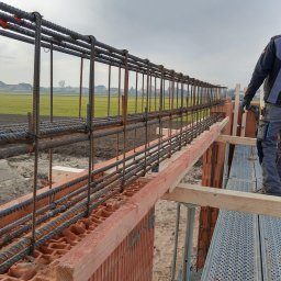 PRAXTON Spółka z o.o. - Zbrojenie ściany z pustaków ceramicznych Ytong, widok z rusztowania na pracownika w odzieży roboczej. W tle pole i pochmurne niebo.