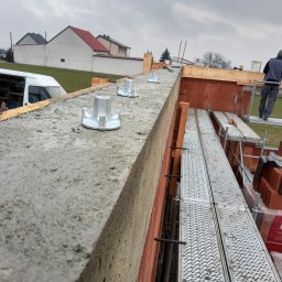 PRAXTON Spółka z o.o. - Wznoszenie muru z pustaków ceramicznych Porotherm. Widoczne metalowe kotwy w betonie i rusztowanie z pracownikiem. Budowa domu w trakcie realizacji.