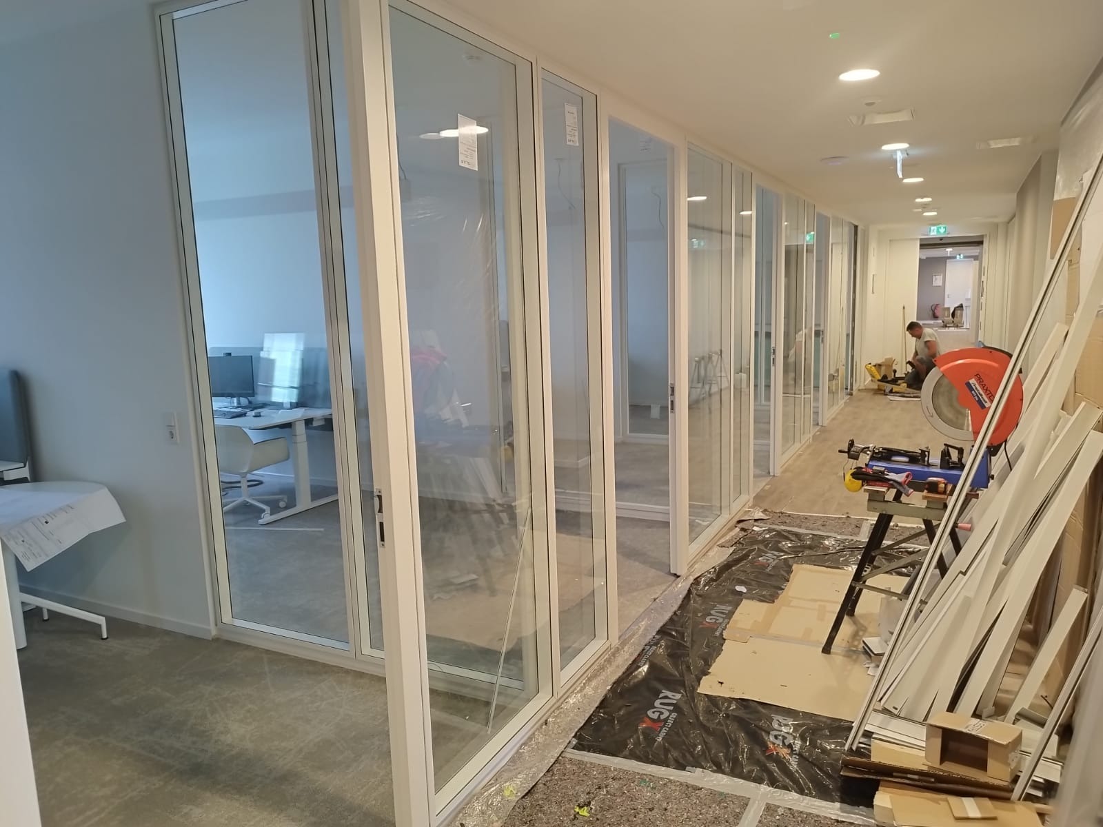 Montaż szklanych ścianek działowych w biurze. Widoczne narzędzia i materiały budowlane, pracownik w oddali. Białe ramy, przezroczyste szkło.
