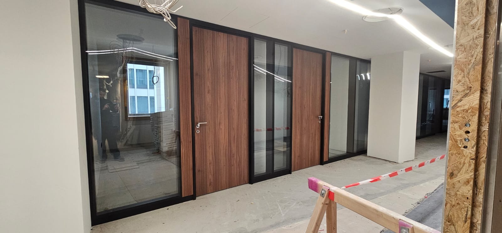Wnętrze biura w trakcie remontu: szklane ścianki działowe z drewnianymi drzwiami, widoczne kable i zabezpieczenia, odblask osoby w szybie.