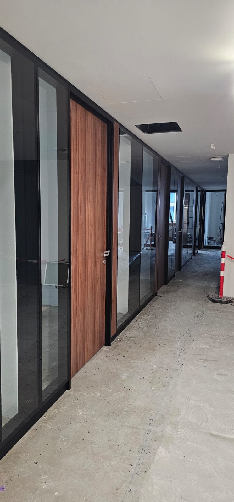 Korytarz biurowy z przeszklonymi ścianami działowymi i drzwiami z okleiną drewnopodobną w czarnych ramach. Widoczne niedokończone podłoże podłogowe.