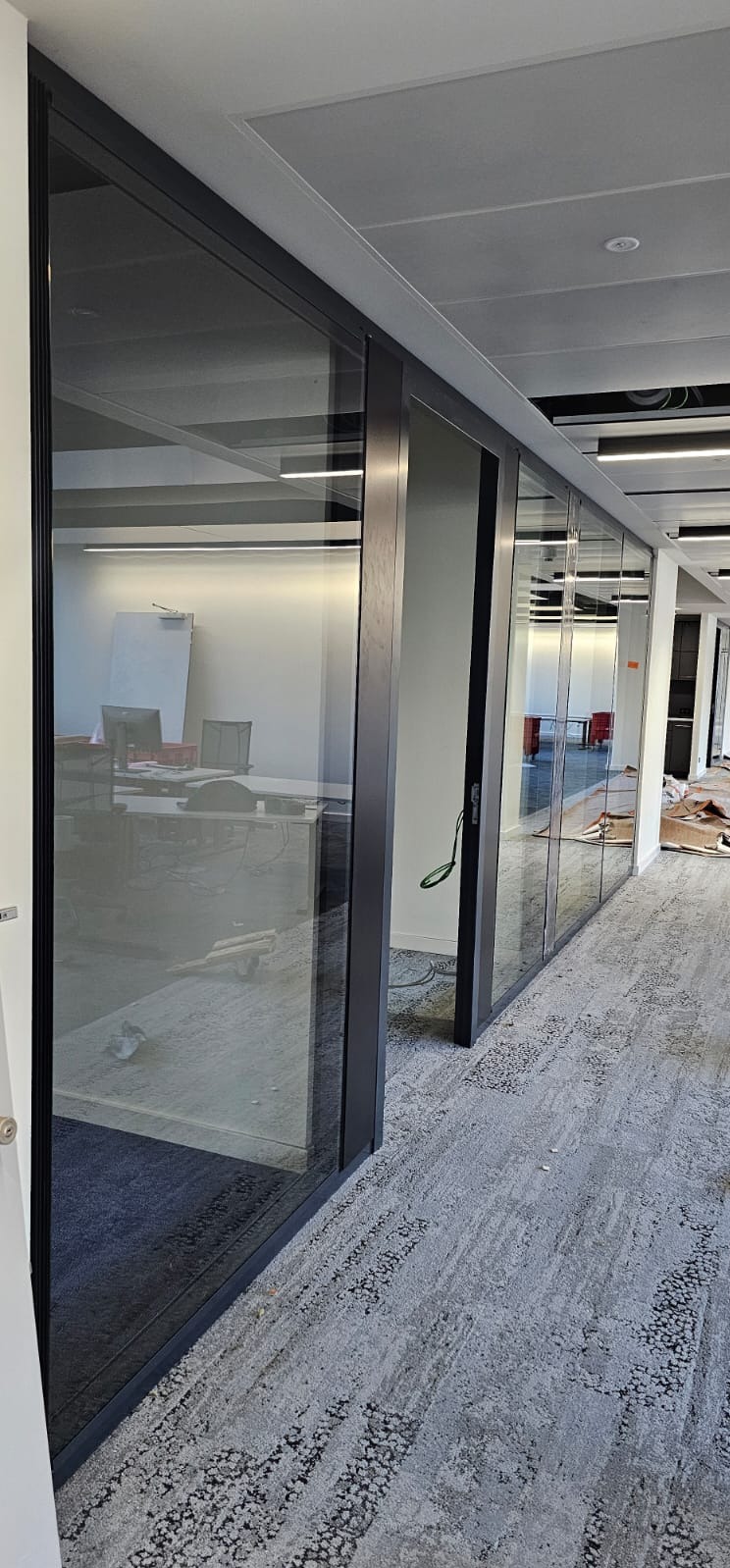 Przeszklone ściany działowe z ciemną ramą w biurze, widok na wnętrze z biurkami i krzesłami. Na podłodze szara wykładzina dywanowa.