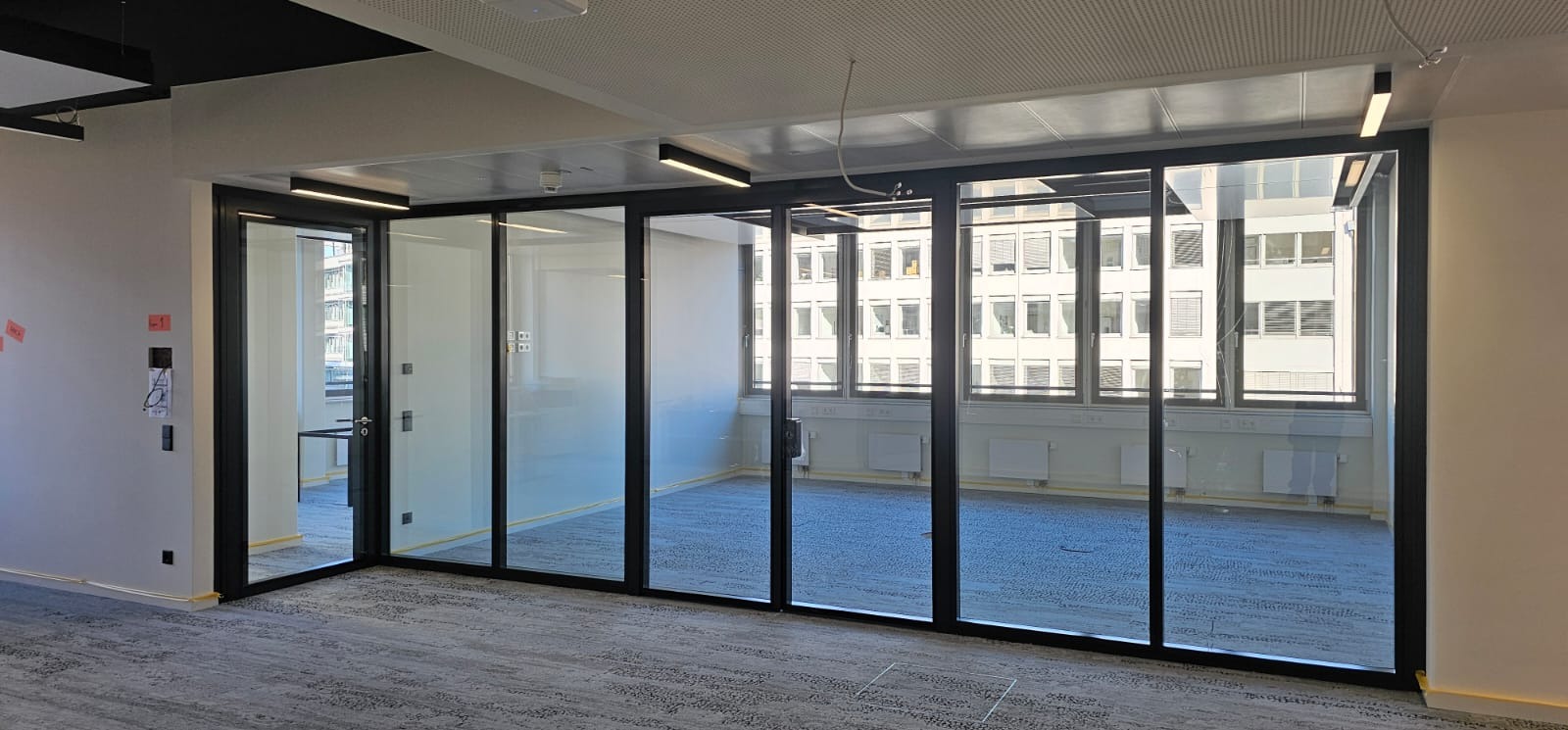 Przeszklona ściana działowa z czarnymi ramami w biurze. Widok na budynek z oknami. Szara wykładzina podłogowa. Lampy sufitowe. Nowoczesny design.