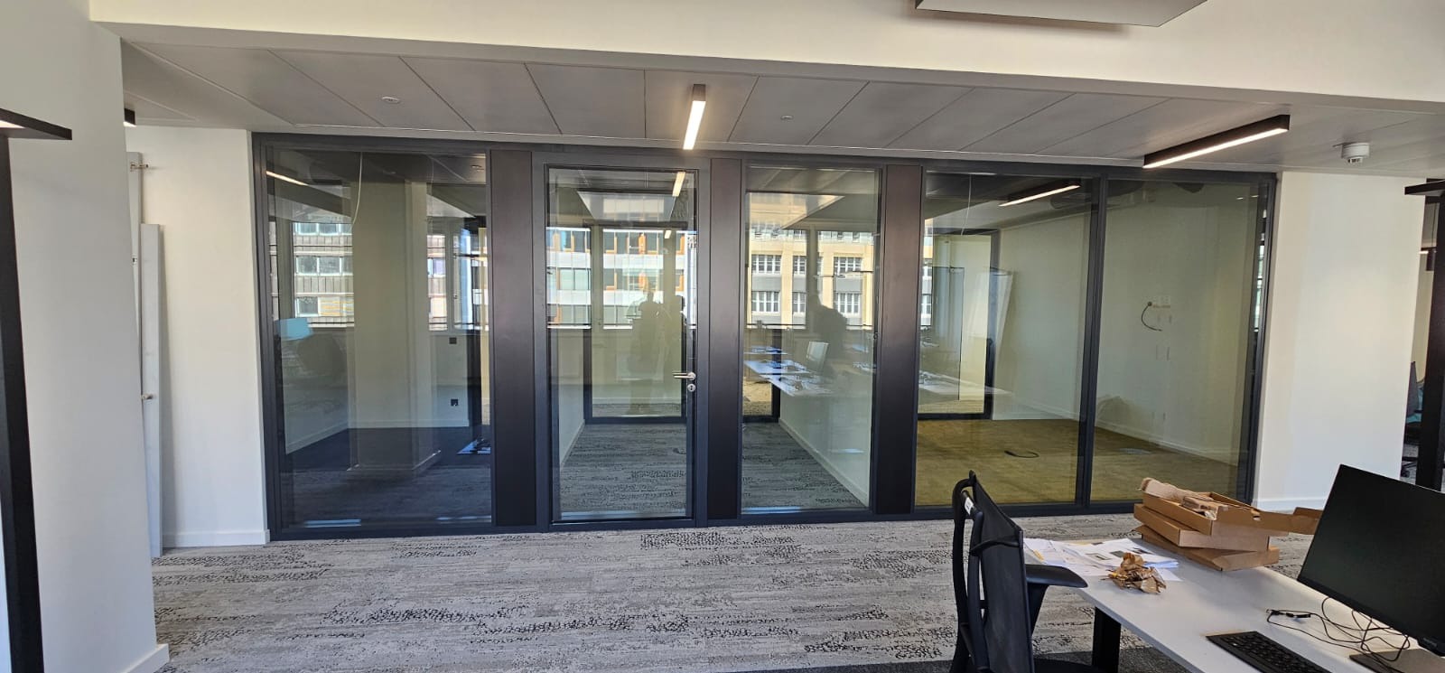 Nowoczesne, przeszklone ścianki działowe w biurze z ciemnymi ramami, widok z korytarza, zza biurka z komputerem i kartonami. W tle budynki odbijające się w szybach.
