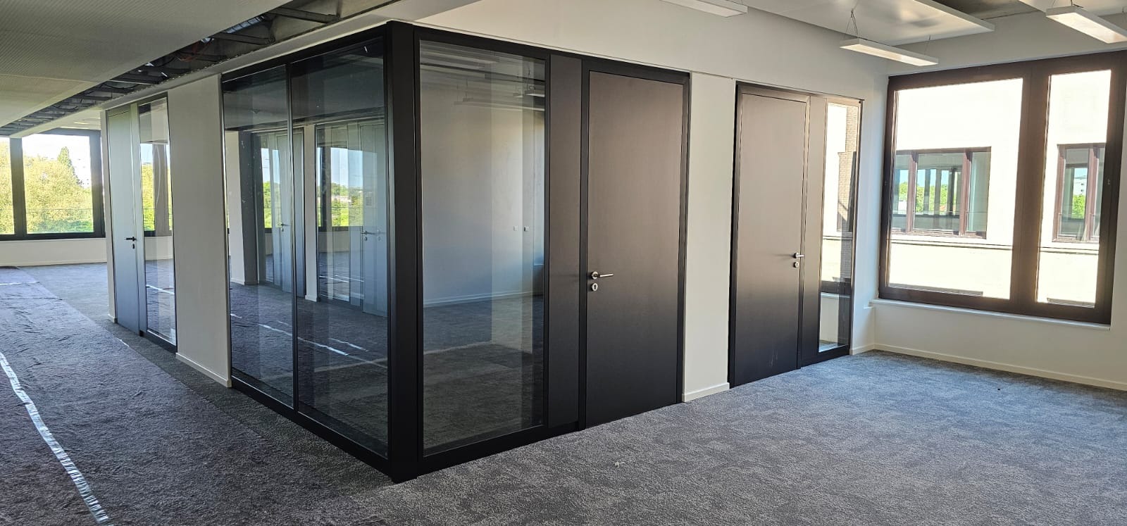 Nowoczesne biuro po remoncie: szklane ścianki działowe z czarnymi ramami, ciemne drzwi i duże okna z widokiem na zieleń. Wykładzina dywanowa w szarościach. Oświetlenie sufitowe.