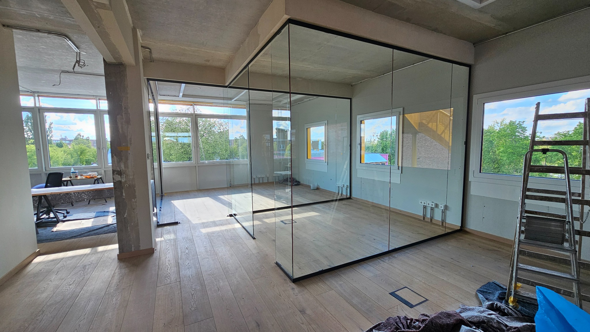 Szklana ścianka działowa w biurze z drewnianą podłogą, widok na zieleń za oknami. Ścianka z czarnymi ramami, drabina oparta o ścianę z oknem.