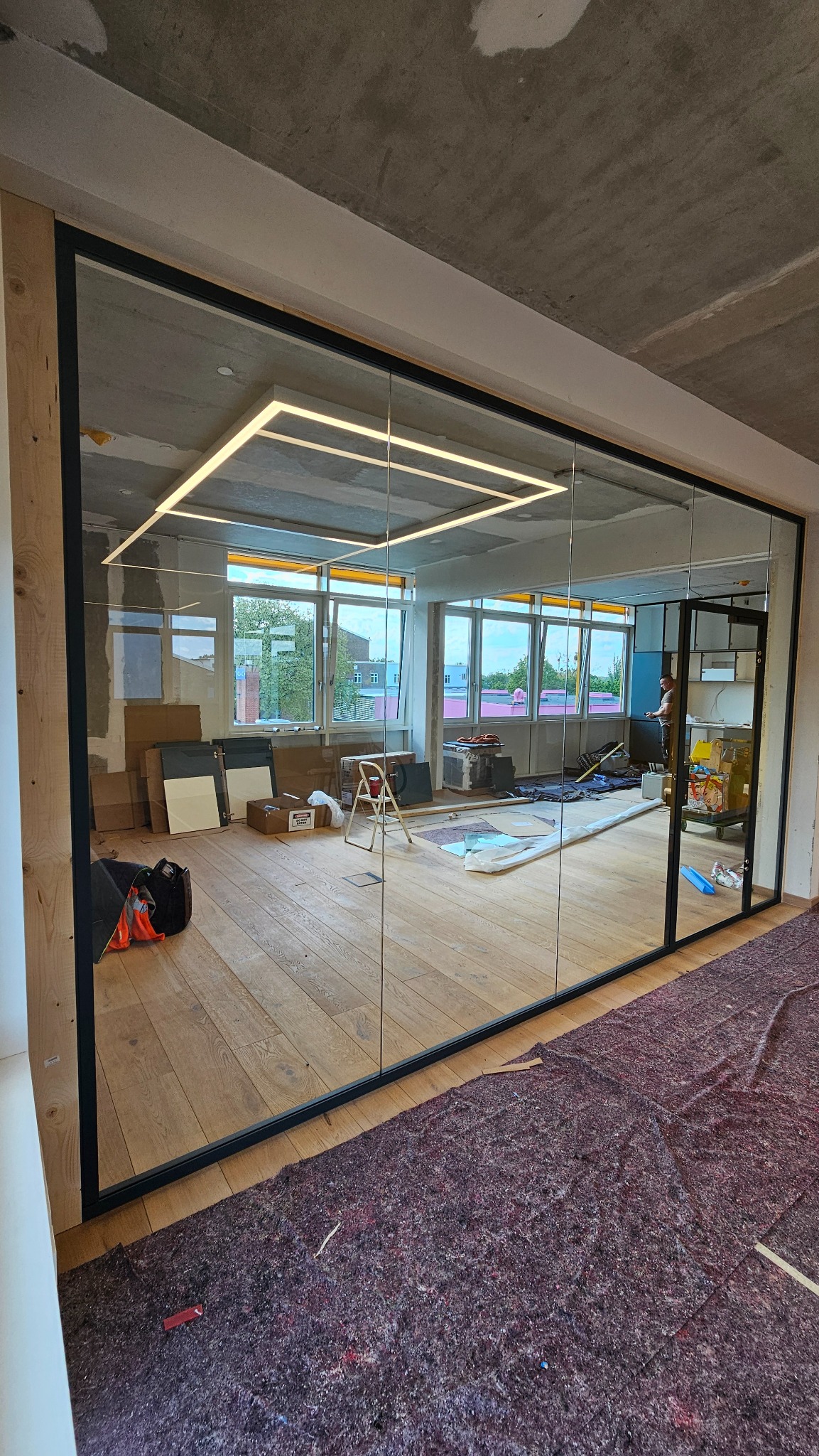 Szklana ściana działowa z czarnymi profilami w biurze, widoczne okna i drewniana podłoga. Oświetlenie sufitowe w kształcie kwadratów. Częściowo zabezpieczona podłoga.