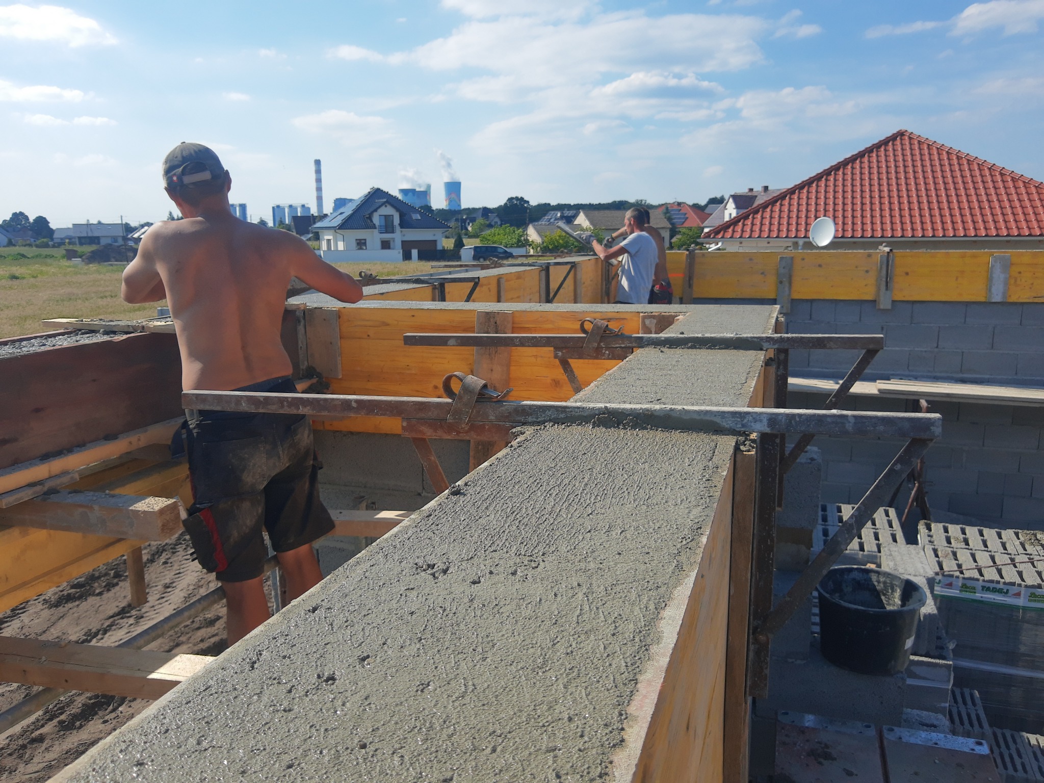 Wylewanie betonowego stropu domu jednorodzinnego. Widoczni dwaj budowlańcy, szalunki, bloczki betonu komórkowego i elementy zbrojenia. W tle zabudowa jednorodzinna.