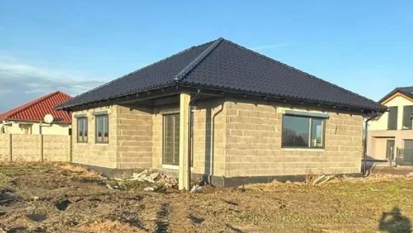 Niedokończony dom jednorodzinny z szarego bloczka, widoczne okna z roletami zewnętrznymi, czarny dach, w tle inne budynki i błękitne niebo.