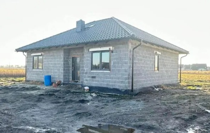 Surowy, parterowy dom z szarego bloczka, z oknami z roletami zewnętrznymi, w trakcie budowy, na tle pola. Widoczny niebieski pojemnik i narzędzia.