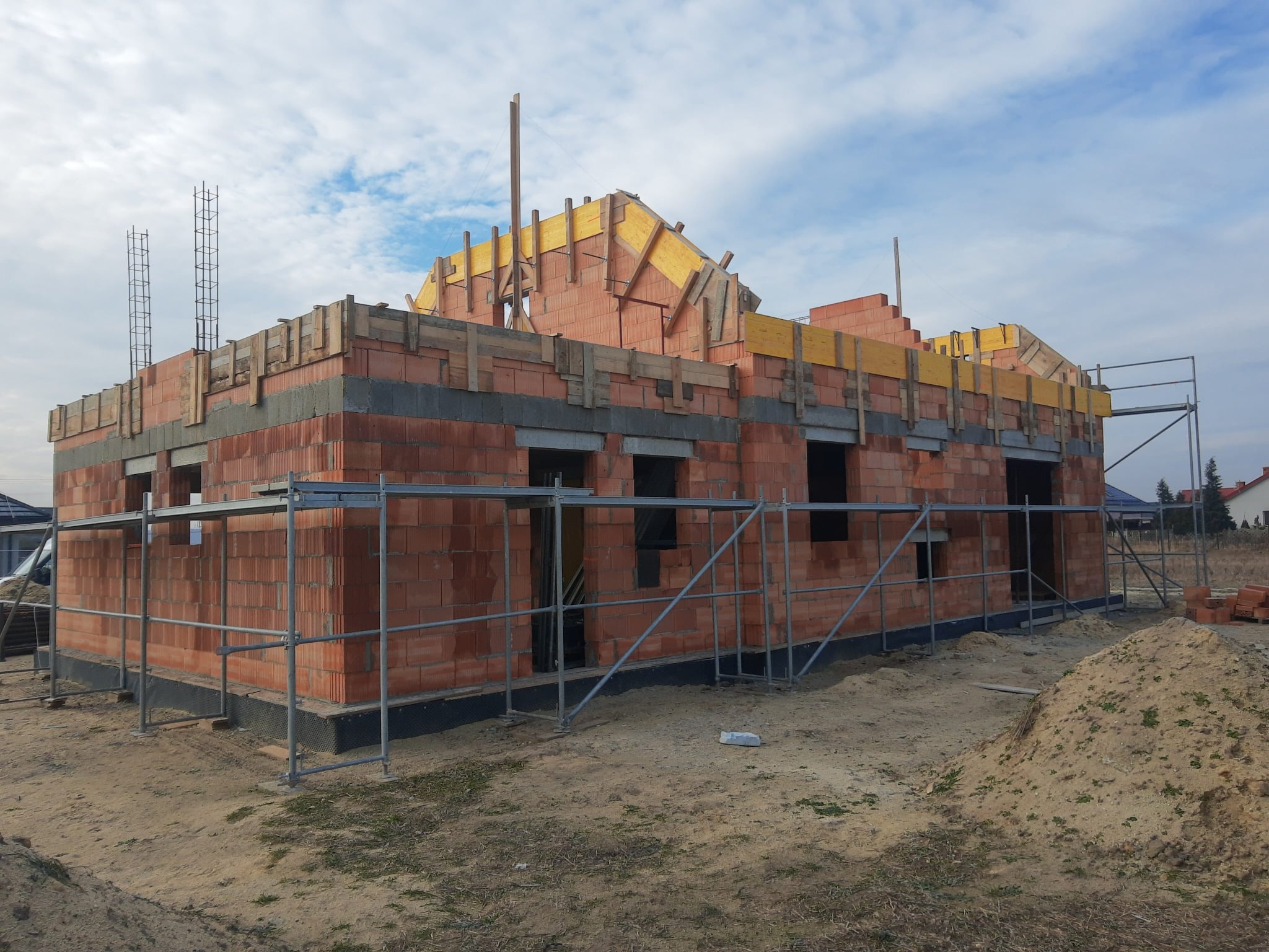 Budowa domu z czerwonej cegły, widoczne rusztowania i elementy konstrukcji dachu. Częściowo wzniesione ściany i przygotowane otwory okienne.