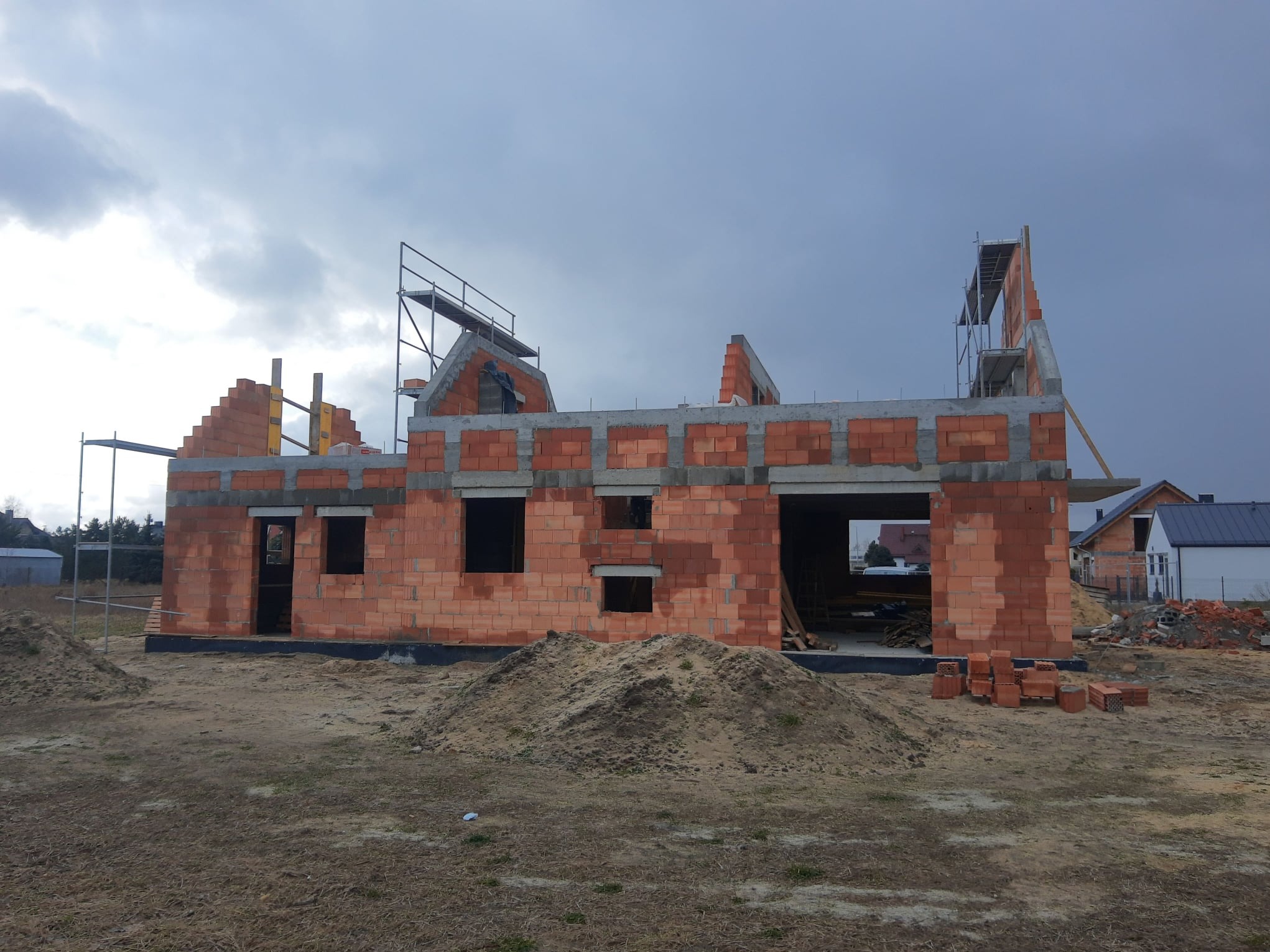 Budowa domu jednorodzinnego w trakcie wznoszenia ścian z czerwonej cegły. Widoczne rusztowania i przygotowany teren. Pochmurne niebo w tle, sąsiednie budynki.