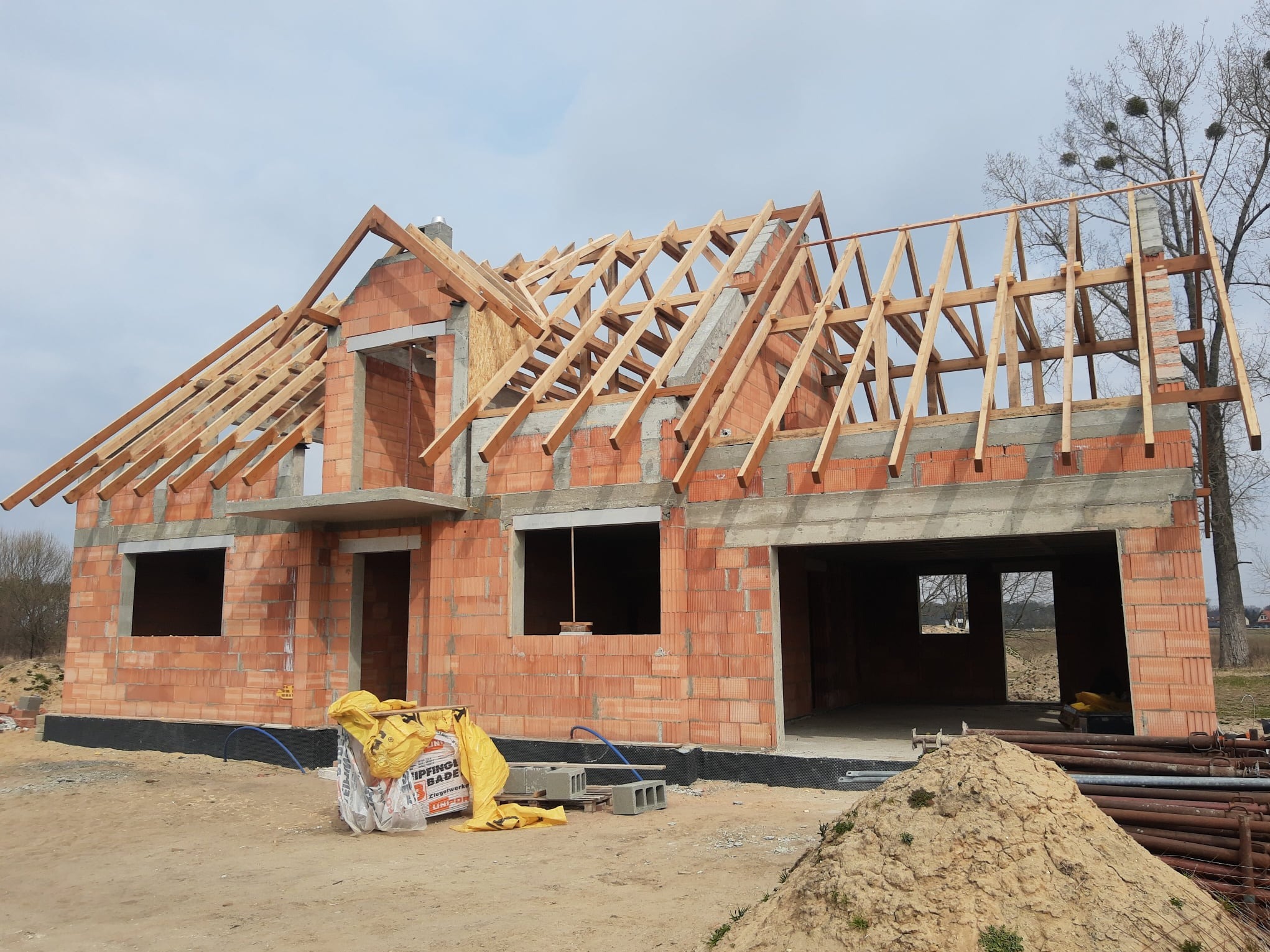 Surowy stan domu z czerwonej cegły z drewnianą konstrukcją dachu w trakcie budowy. Widoczne pustaki, betonowe nadproża i elementy konstrukcyjne dachu.