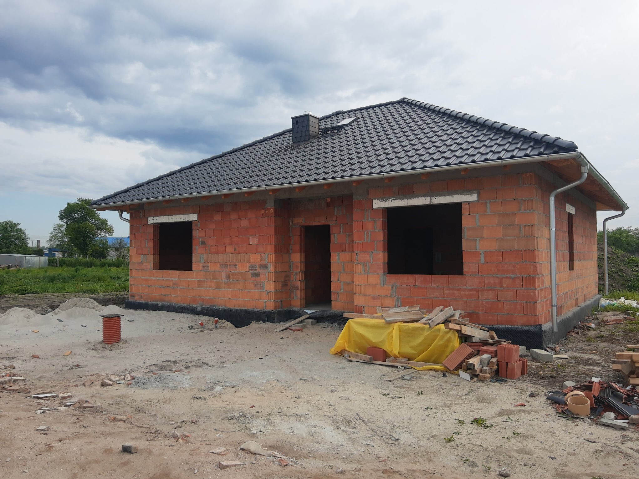 Surowy, parterowy dom z czerwonej cegły z czarnym dachem pod pochmurnym niebem. Widoczne otwory okienne i drzwiowe, teren wokół domu w trakcie budowy, materiały budowlane.