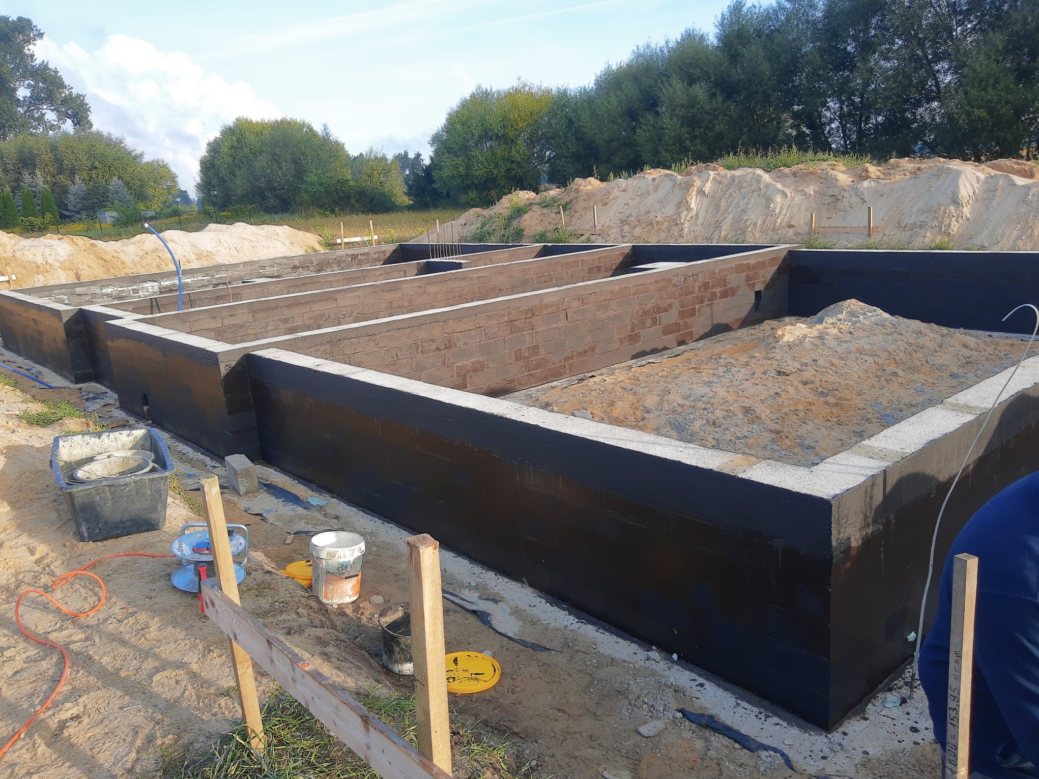 Zabezpieczone fundamenty domu z bloczków betonowych, wypełnione piaskiem, z widocznymi narzędziami budowlanymi i otoczeniem placu budowy w słoneczny dzień.