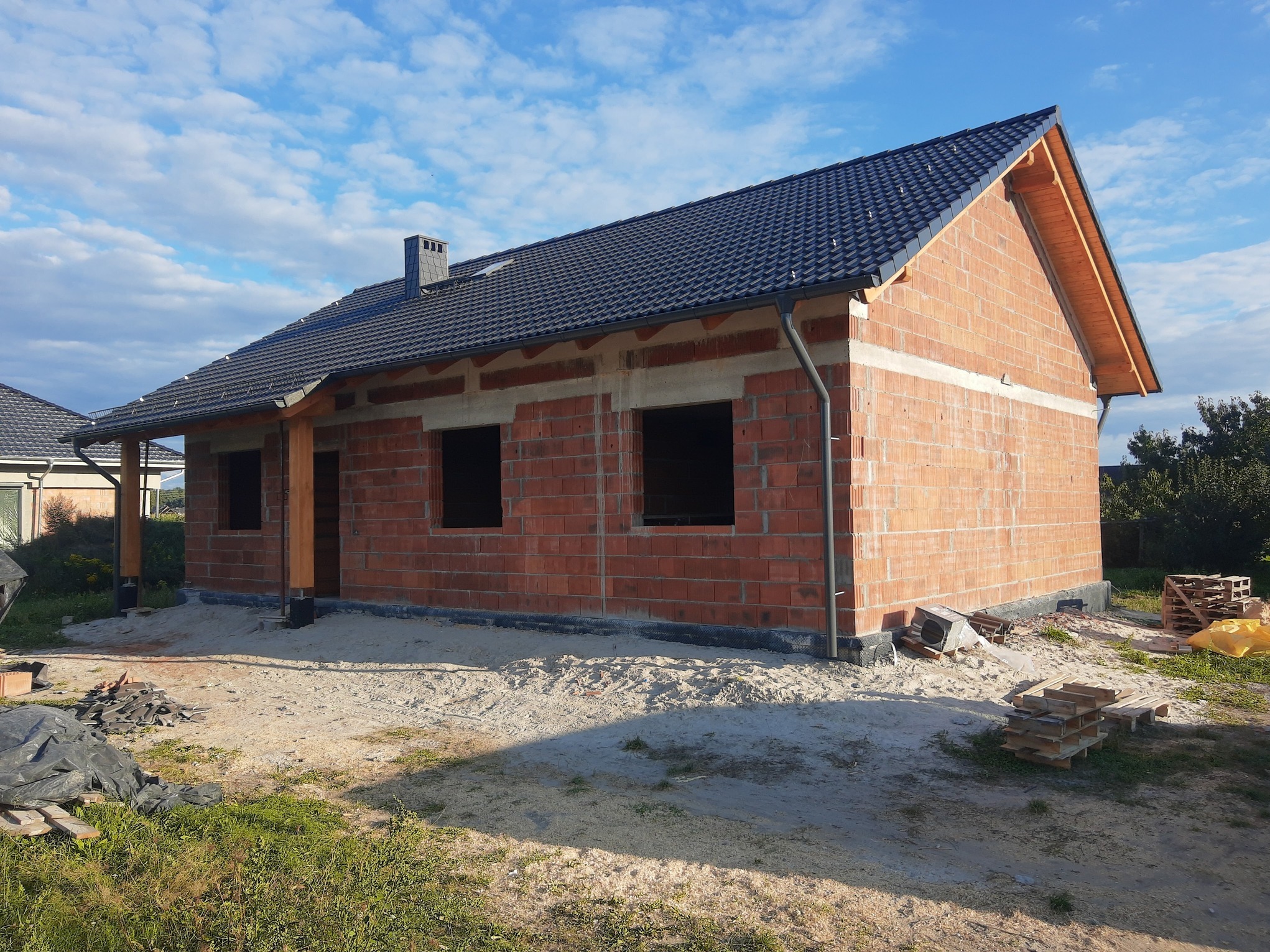 Niedokończony dom z czerwonej cegły pod błękitnym niebem, z widocznym dachem z czarnej dachówki i drewnianą konstrukcją podbitki. Wokół piasek i palety.