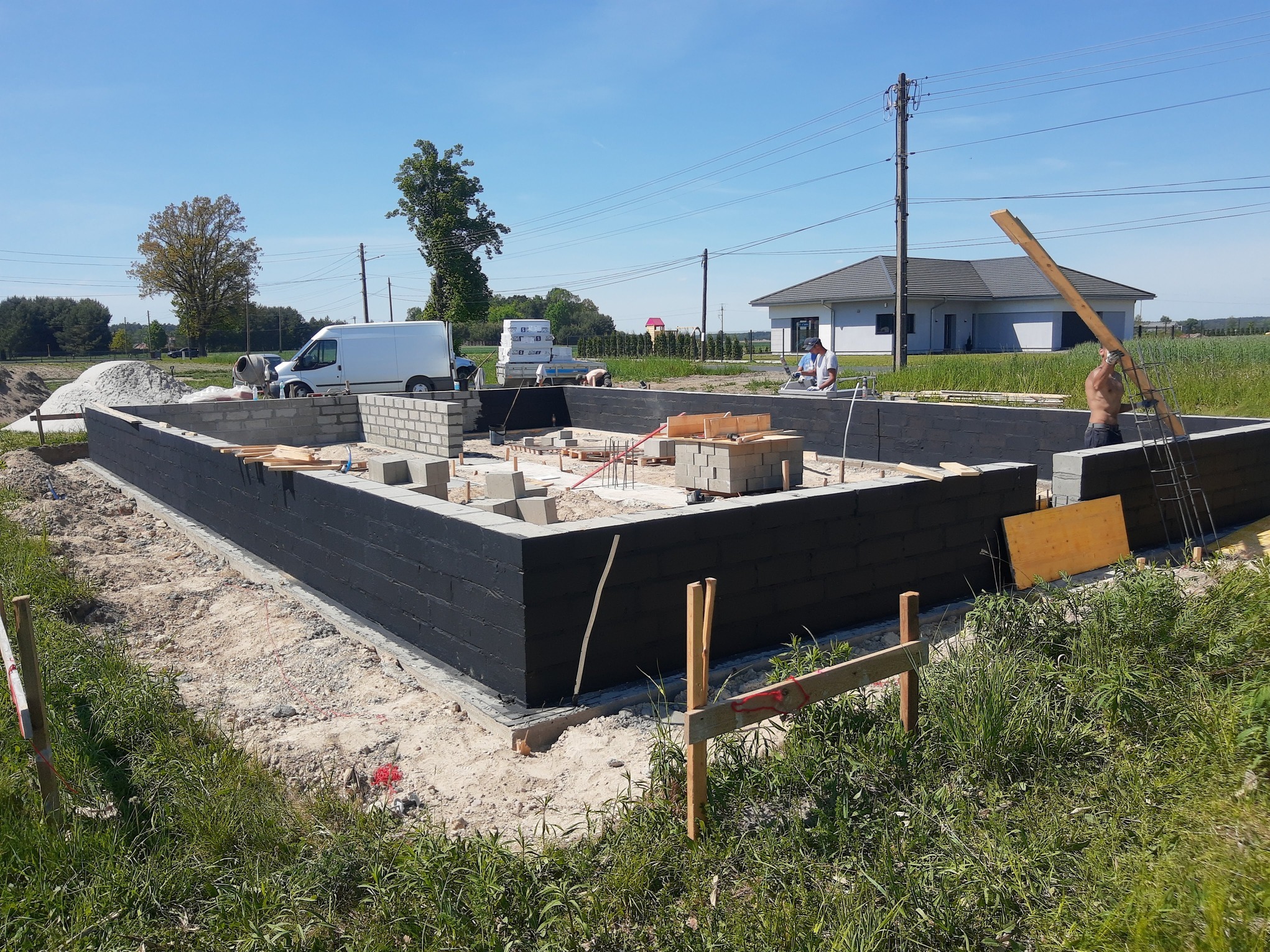 Wznoszenie fundamentów domu jednorodzinnego: murowane ściany z izolacją, pracownicy w trakcie robót, widoczny sprzęt budowlany i biały van w tle.