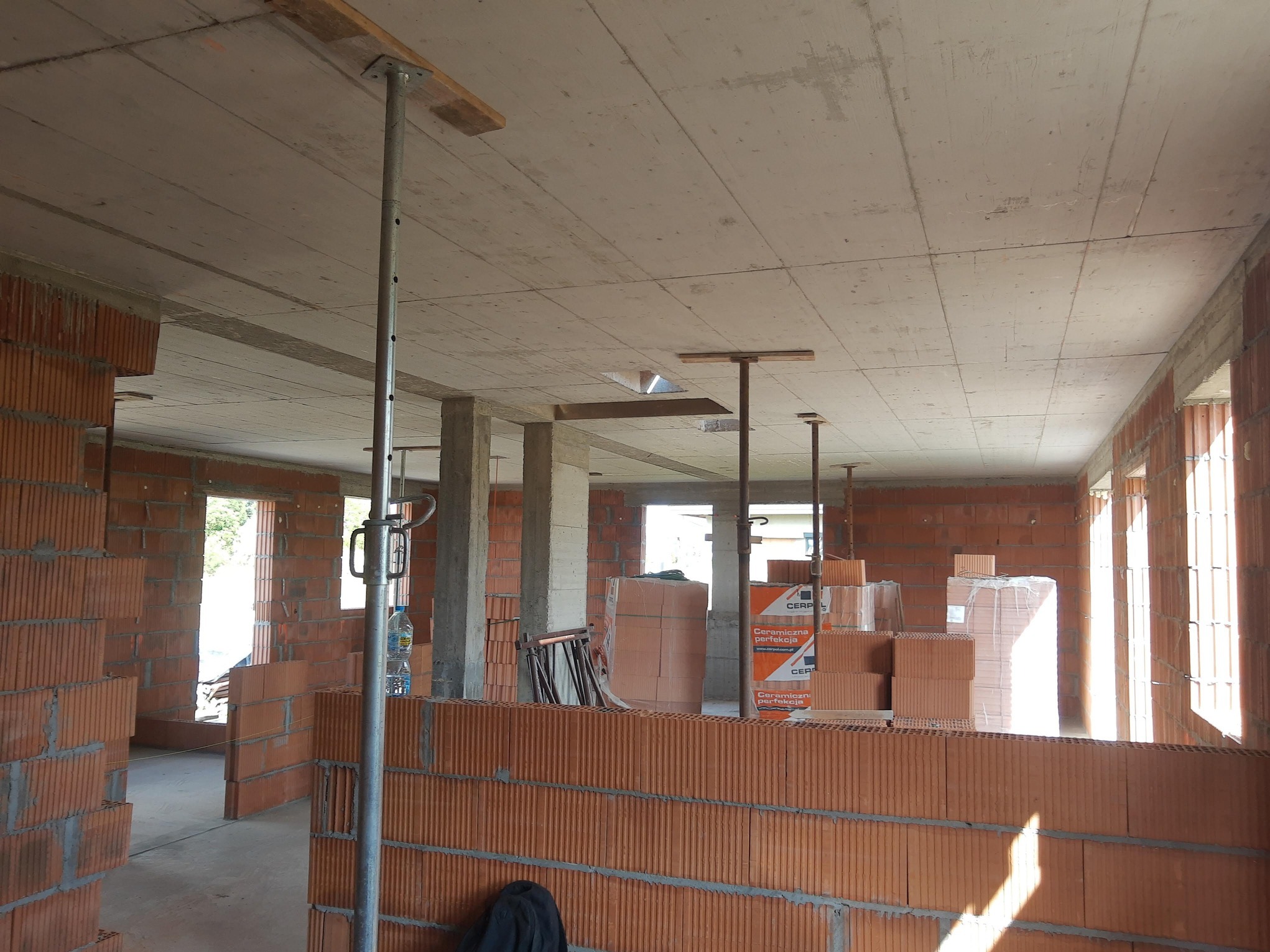 Wnętrze domu w trakcie budowy: ściany z czerwonej cegły, betonowy strop podparty metalowymi stemplami, okna bez ram. Widoczne materiały budowlane.