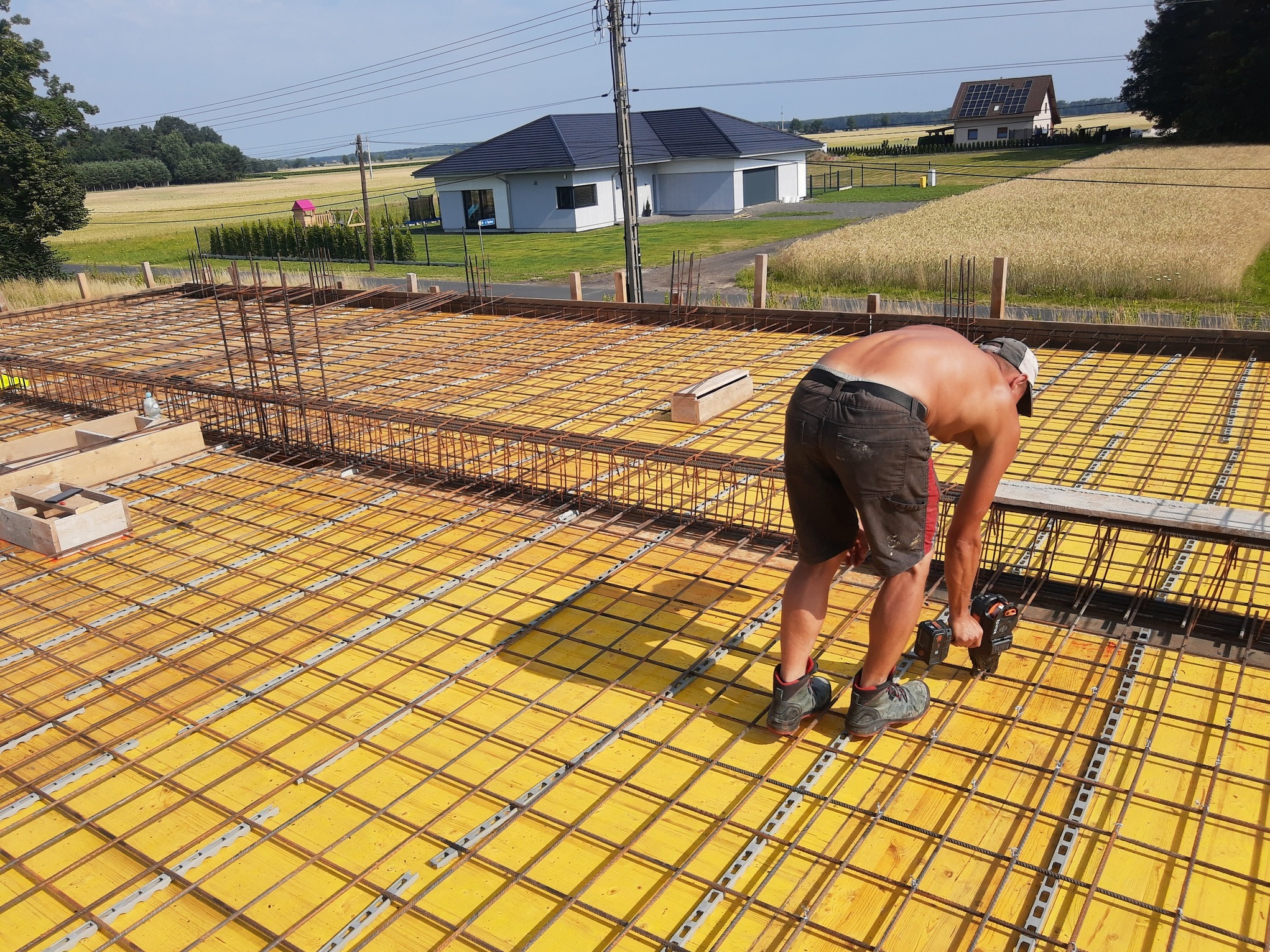 Prace zbrojarskie na budowie domu jednorodzinnego: zbrojarz łączy stalowe pręty na żółtym szalunku, w tle dom z ciemną dachówką i krajobraz wiejski.