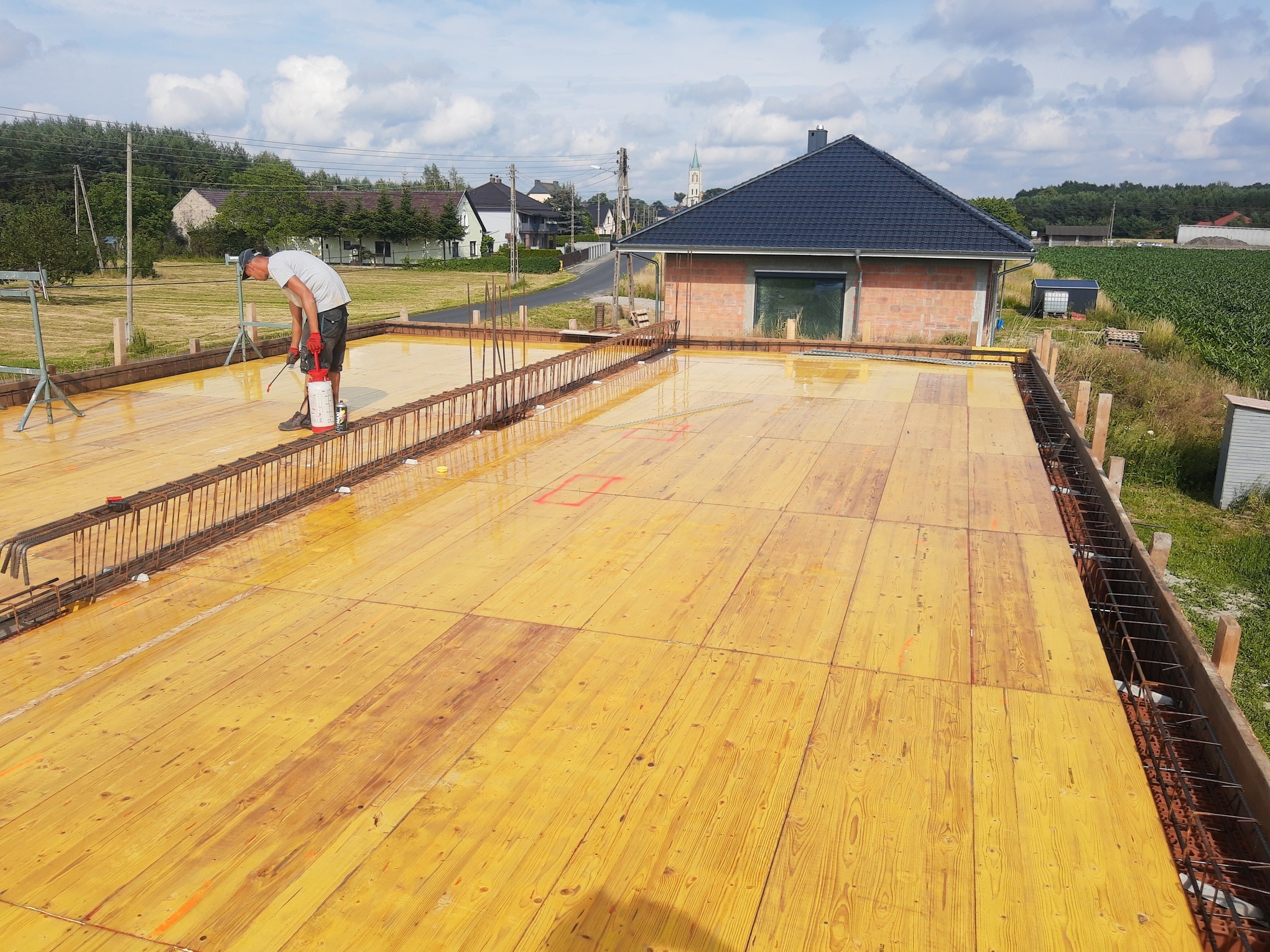 Przygotowanie szalunku i zbrojenia pod fundament domu jednorodzinnego. Widoczny pracownik impregnujący drewniane płyty szalunkowe. W tle zabudowa jednorodzinna i pola uprawne.