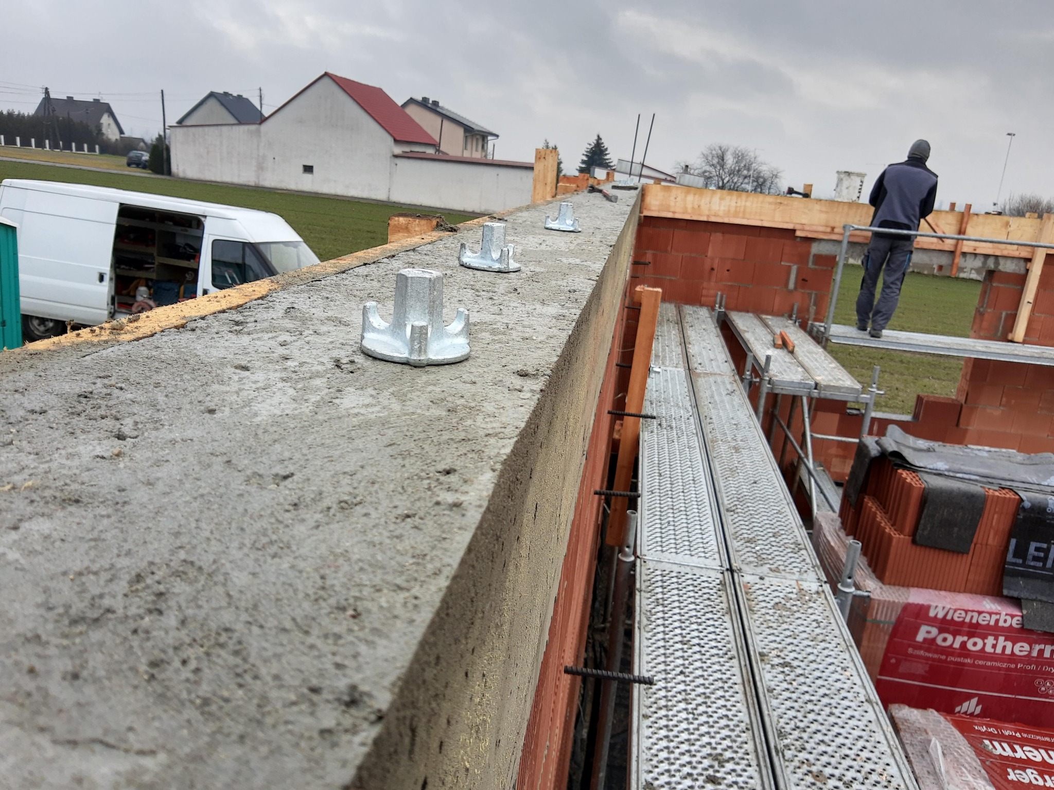 Wznoszenie muru z pustaków ceramicznych Porotherm. Widoczne metalowe kotwy w betonie i rusztowanie z pracownikiem. Budowa domu w trakcie realizacji.