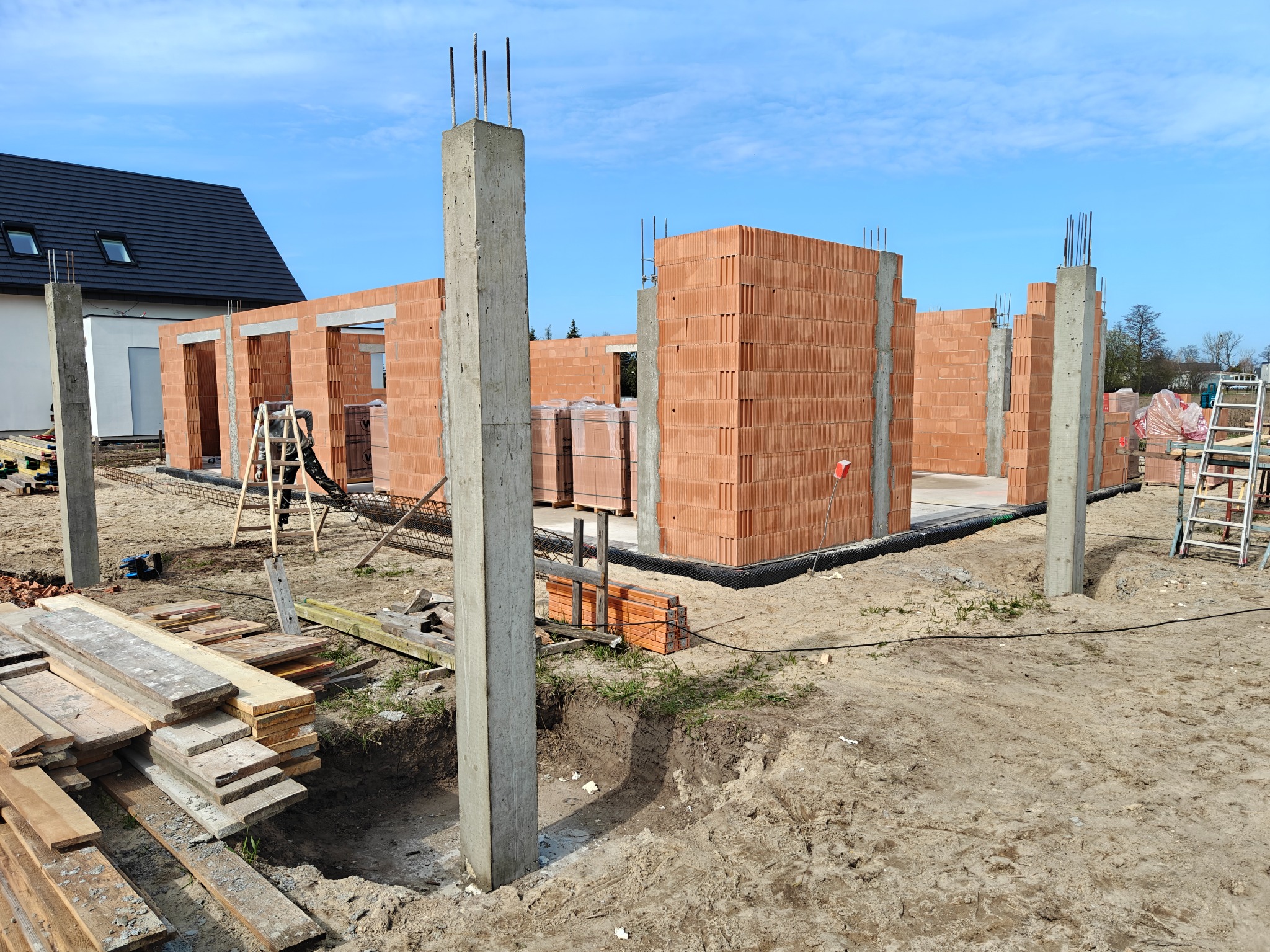 Wznoszenie ścian domu z czerwonej cegły ceramicznej. Widoczne betonowe słupy, drewniane deski i drabina. W tle fragment dachu i błękitne niebo.