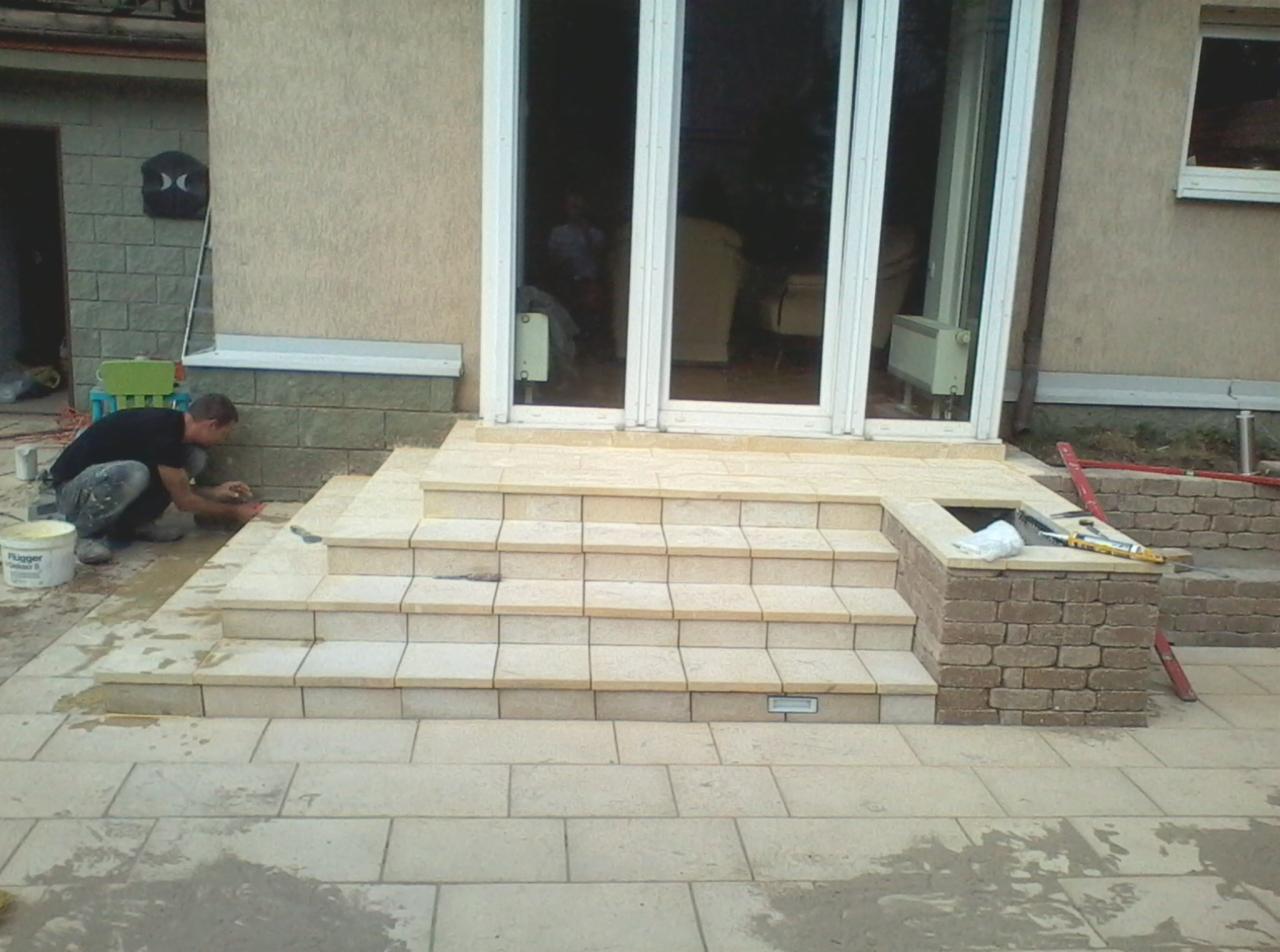 Układanie płytek na schodach wejściowych do domu, pracownik w trakcie fugowania, widoczny kubeł z zaprawą i narzędzia, jasne płytki i kostka brukowa.