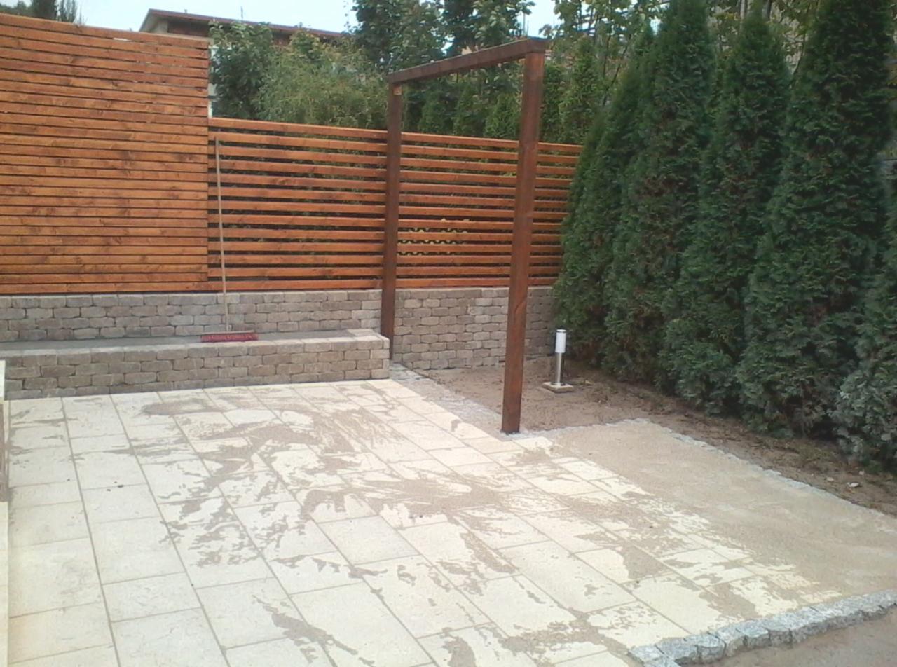 Nowo wyłożony taras z jasnych płyt, drewniany płot z poziomych desek na kamiennej podmurówce oraz drewniana pergola w ogrodzie z tujami.