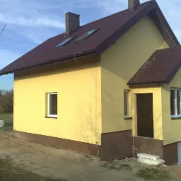 Żółty dom z brązowym dachem i podmurówką, widok z boku, z widocznym fragmentem terenu budowy i nieułożonymi płytami chodnikowymi.