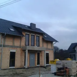 Dwukondygnacyjny dom w trakcie budowy z bloczków, widoczne okna, dach z dachówki ceramicznej Roben, drugi dom w tle, pochmurne niebo.