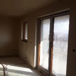 Pokój w stanie deweloperskim z oknami balkonowymi, ściany przygotowane pod malowanie, widoczne instalacje grzewcze w podłodze.