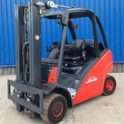 Czerwono-szary wózek widłowy Linde H25T z widocznym znakiem ostrzegawczym na maszcie, stojący na betonowej powierzchni przed niebieską blachą falistą.