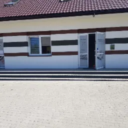 Elewacja budynku biurowego z dwoma wejściami, na drzwiach godziny otwarcia, białe ściany z brązowymi pasami, szary chodnik z kostki brukowej.