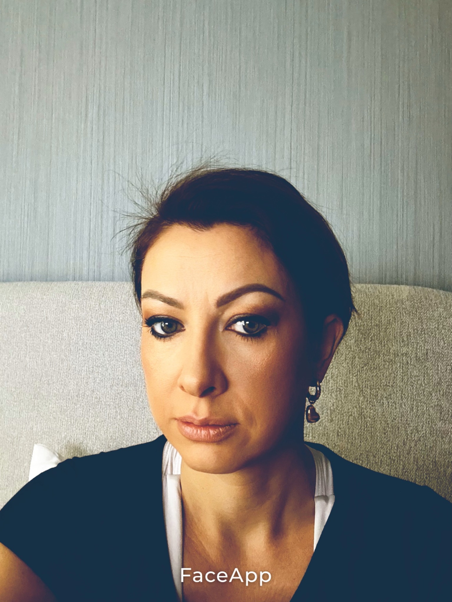 Portret kobiety z krótkimi, ciemnymi włosami i delikatnym makijażem, ubranej w ciemną bluzkę z białym topem. W tle jasnoszara ściana i fragment sofy.