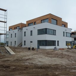 MAXI-BUD USŁUGI OGÓLNOBUDOWLANE MASZOTA HENRYK - Nowoczesny, dwukondygnacyjny budynek z białymi ścianami i drewnianą elewacją na poddaszu, w trakcie budowy, z widocznym sprzętem budowlanym i rusztowaniami.