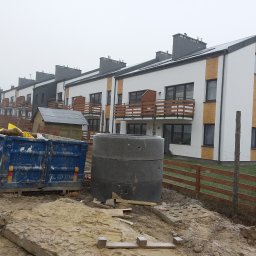 MAXI-BUD USŁUGI OGÓLNOBUDOWLANE MASZOTA HENRYK - Nowoczesne domy szeregowe z balkonami, wykończone białą elewacją z drewnianymi akcentami. Na pierwszym planie kontener i betonowe kręgi na placu budowy.