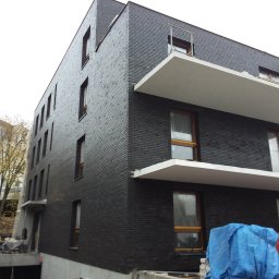 MAXI-BUD USŁUGI OGÓLNOBUDOWLANE MASZOTA HENRYK - Nowoczesny budynek z ciemnej cegły z białymi balkonami, w trakcie budowy. Widoczne rusztowania i materiały budowlane. Ujęcie z dołu, podkreślające geometrię budynku.